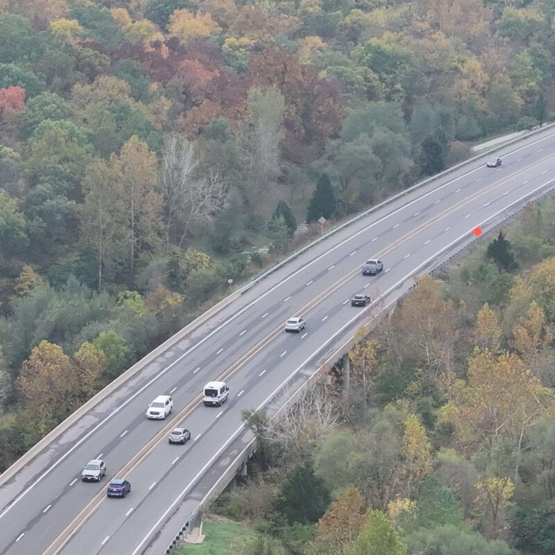 Cars on highway amid dense autumn trees, aerial view.
