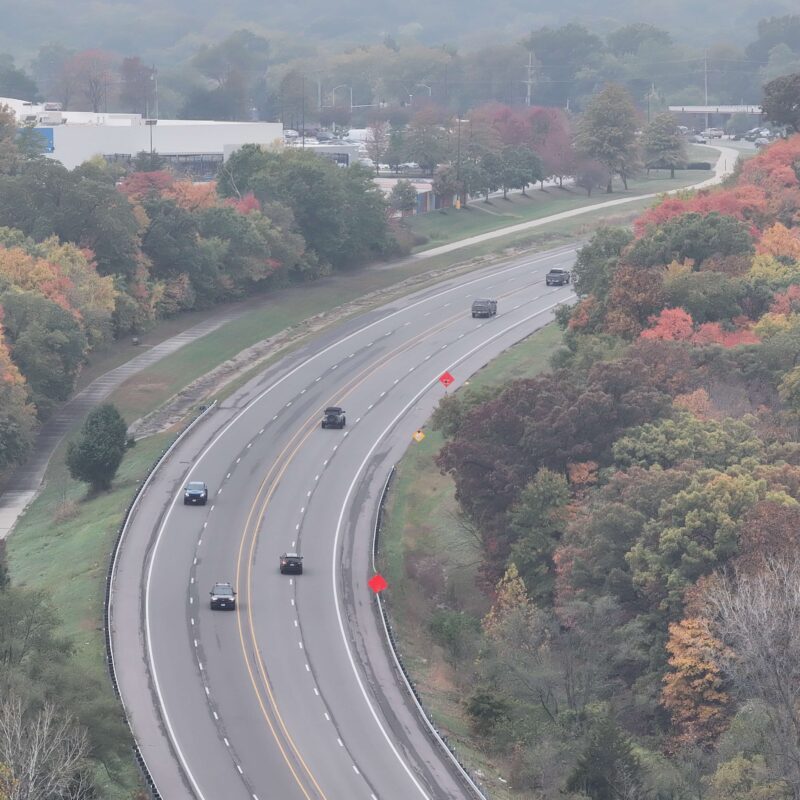 Curved highway with cars, autumn trees, and a sidewalk.