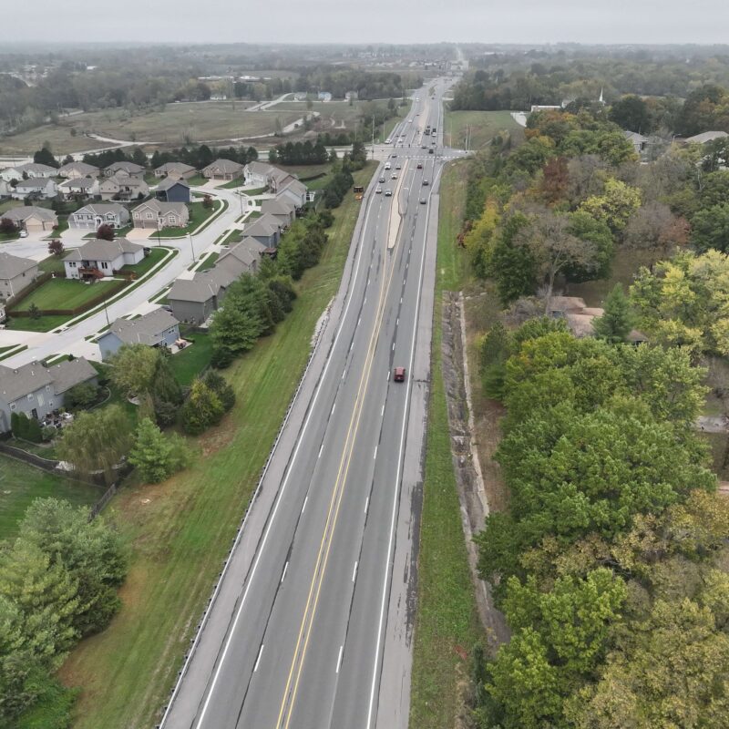 Aerial view of highway, cars, houses left, trees right.
