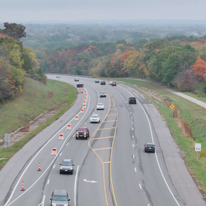 Cars on divided highway, lane closed by cones, autumn trees.