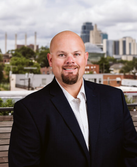 Bald man in suit sits on bench before city skyline.