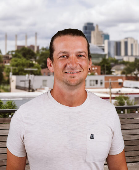 Man with brown hair tied back, light t-shirt, city backdrop.