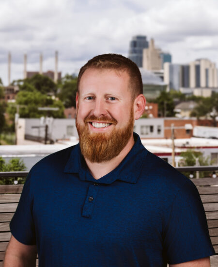 Man with reddish beard smiles outdoors, cityscape behind him.