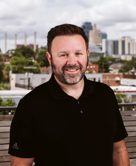 Bearded man in black polo stands outdoors before city skyline.