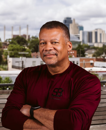 Man in maroon shirt smiles on bench, city skyline behind.