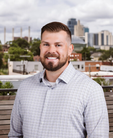Smiling bearded man in plaid shirt, cityscape behind.