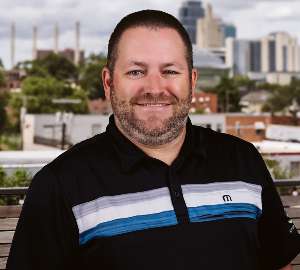 Bearded man in black polo outside, cityscape blurred behind.