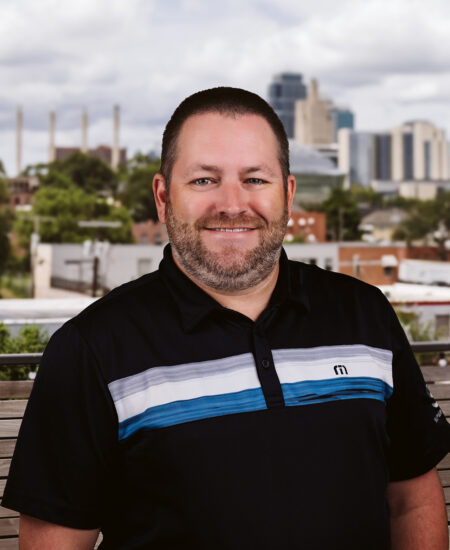 Bearded man in black polo outside, cityscape blurred behind.