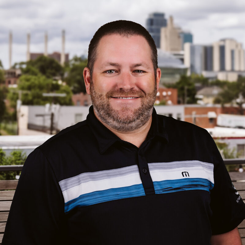 Bearded man in black polo outside, cityscape blurred behind.