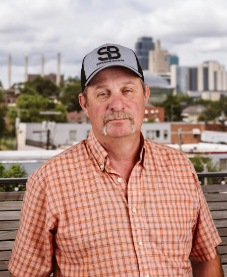 Man in checkered shirt and cap stands outside before cityscape.