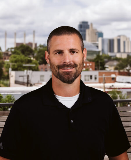 Man with beard in black shirt stands outside by cityscape.