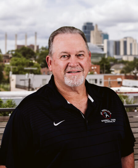 Older man in black polo stands outdoors before cityscape.