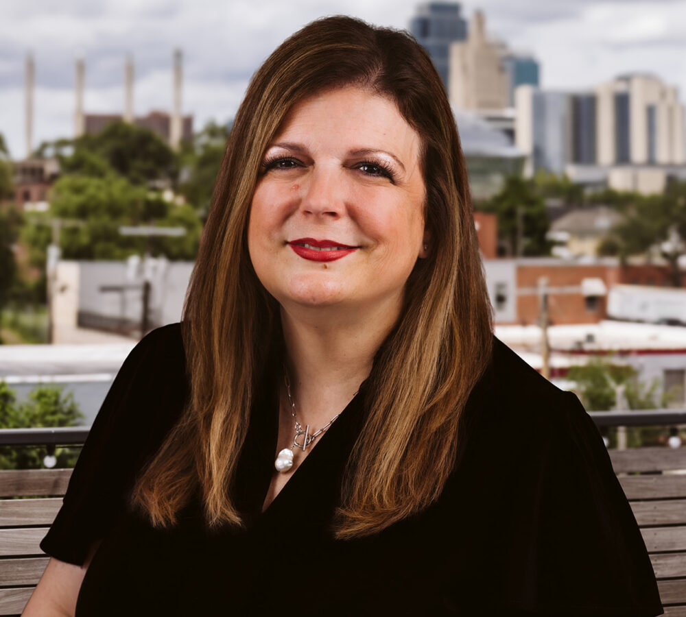 Woman with long brown hair sits on a bench, city behind.