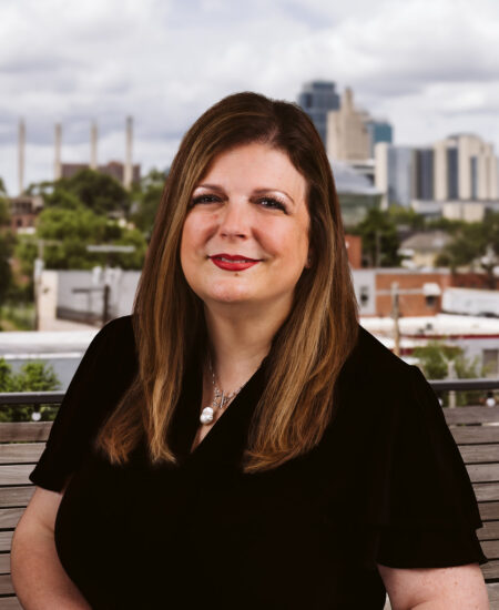Woman with long brown hair sits on a bench, city behind.