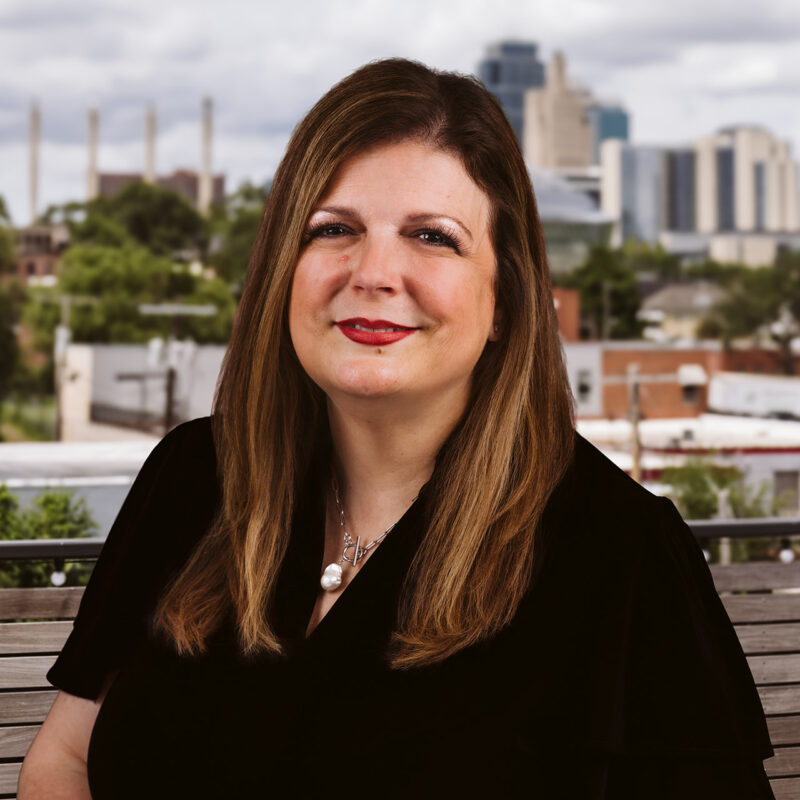Woman with long brown hair sits on a bench, city behind.