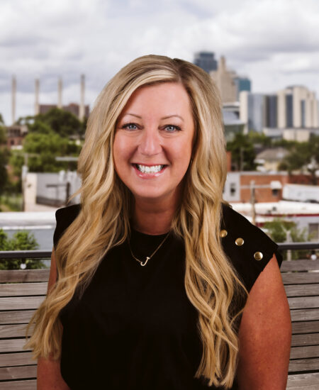 Smiling blonde woman in black top sits on bench cityscape.