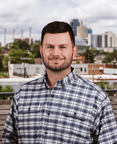 Bearded man in plaid shirt outdoors, cityscape behind him.