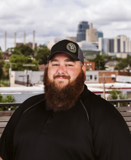 Bearded man in black polo and cap sits outside, city behind.