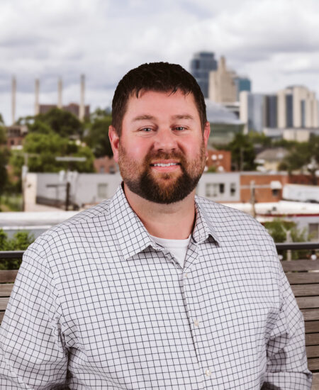 Bearded man in white checkered shirt outdoors, city backdrop.