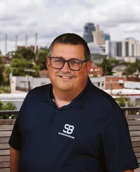 Man in glasses and navy polo sits on outdoor bench, city behind.