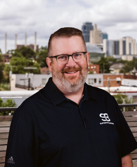 Smiling bearded man with glasses in black shirt, city behind.