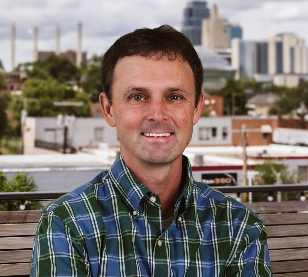 Man in plaid shirt sits on bench, city and clouds behind.