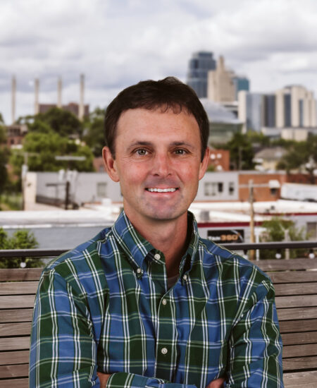 Man in plaid shirt sits on bench, city and clouds behind.