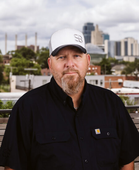 Bearded man in white cap, black shirt, city and clouds behind.