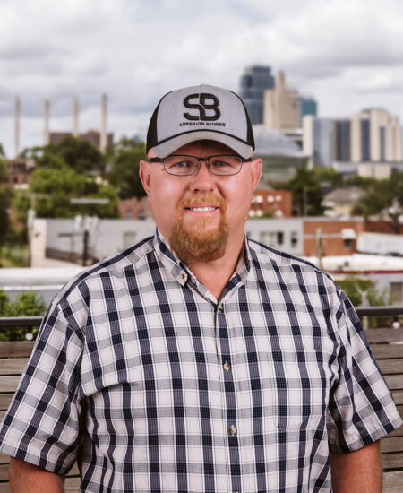 Man in glasses, plaid shirt, and hat outdoors by city.
