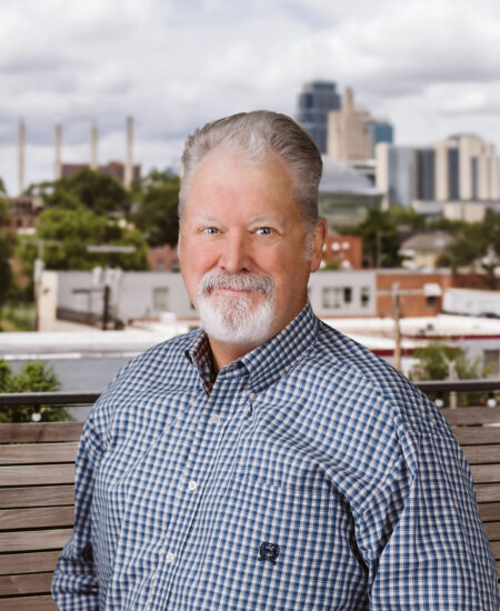 Gray-haired man in blue checkered shirt on bench, city behind.
