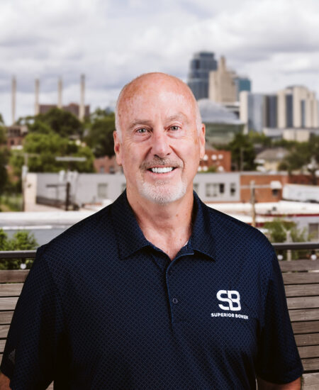 Smiling bald man in navy polo outdoors, city behind.