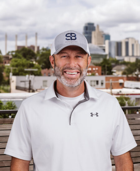 Man in gray cap and white polo smiles outdoors, city behind.