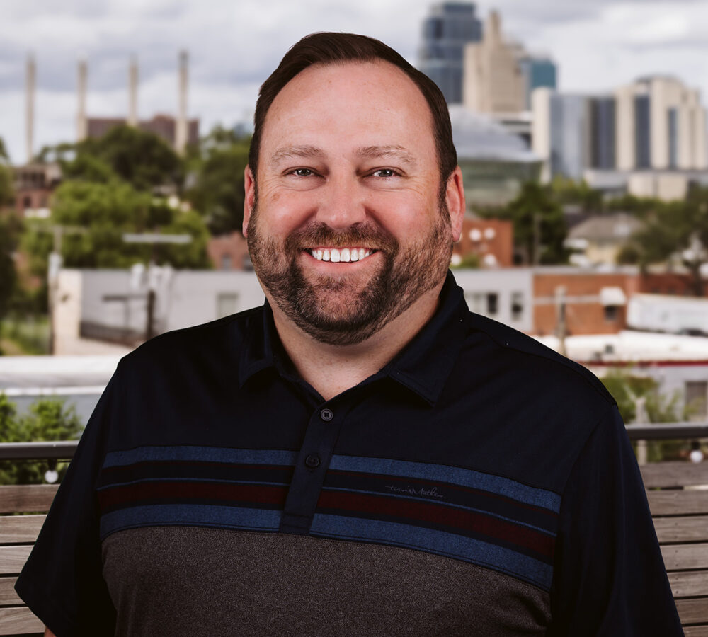 Smiling bearded man outdoors with cityscape and cloudy sky.