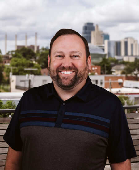 Smiling bearded man outdoors with cityscape and cloudy sky.