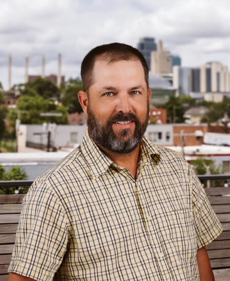 Bearded man in yellow plaid shirt on bench, city behind.