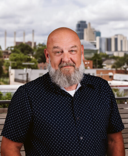 Bald man with gray beard in patterned shirt, city skyline behind.
