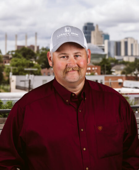 Man with mustache in gray cap, maroon shirt, city background.