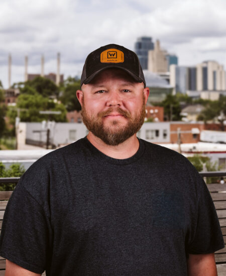 Bearded man in baseball cap outdoors, city and clouds behind.