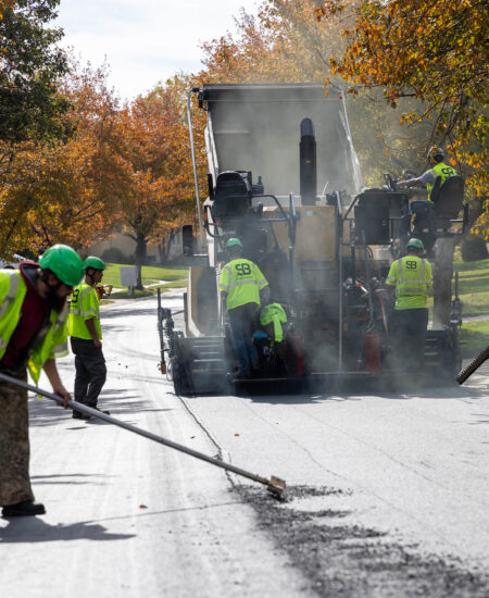 Workers in safety vests repave a street with an asphalt machine.