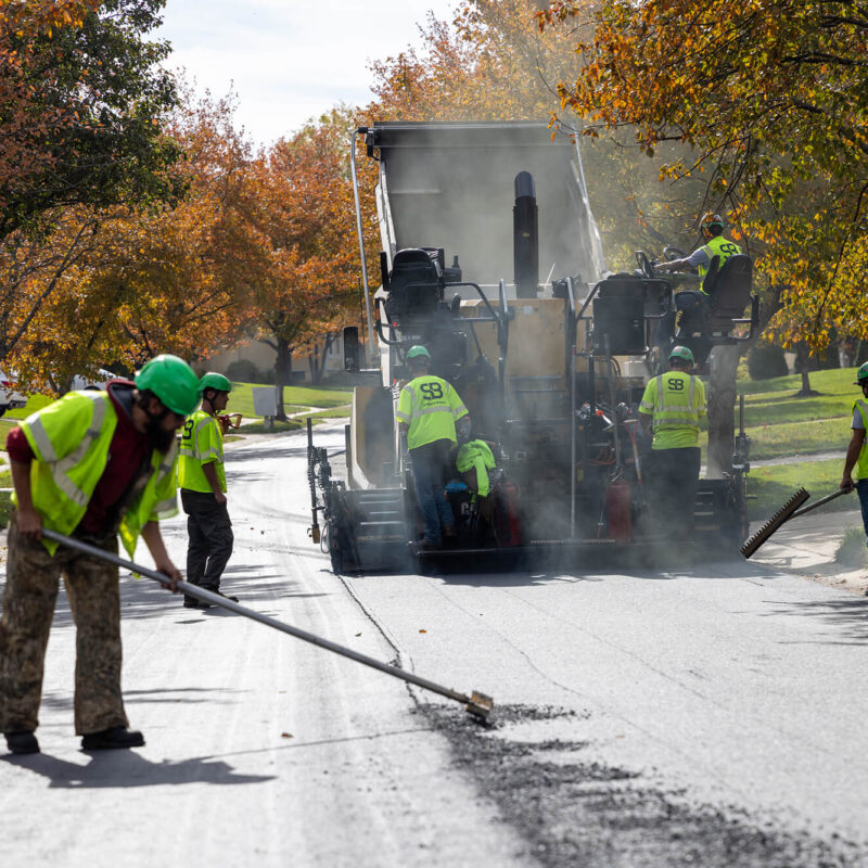 Workers in safety vests repave a street with an asphalt machine.