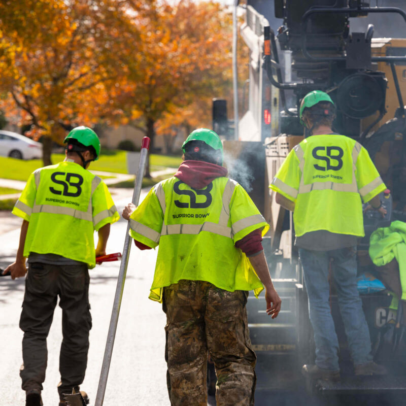 Three workers in safety gear use paving equipment on a road.