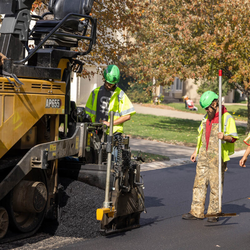 Workers in safety gear pave asphalt on a leafy residential street.