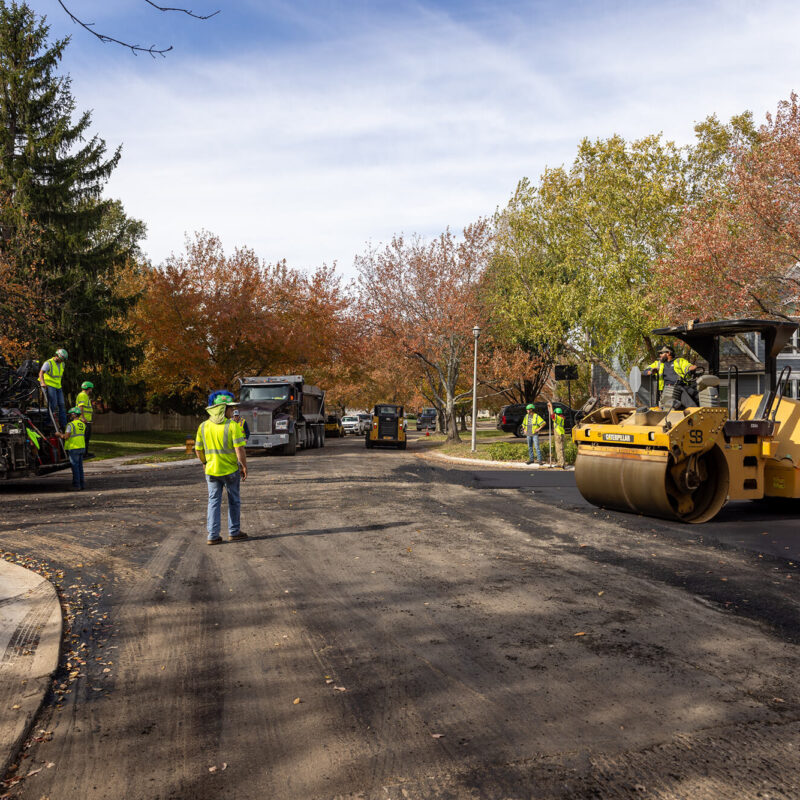 Workers repave street with asphalt paver, steamroller, and truck.
