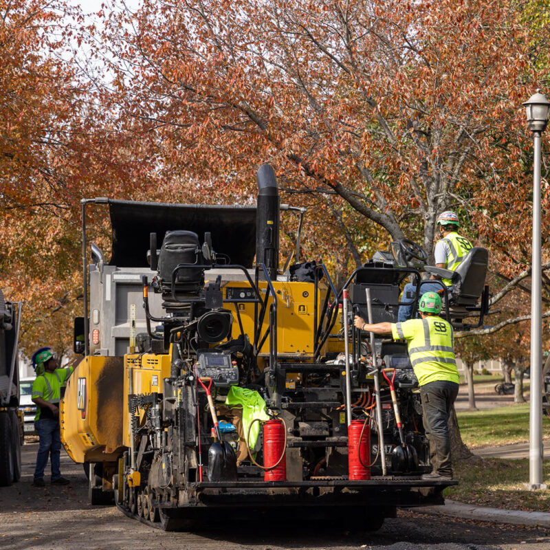 Workers run yellow paving machine on leafy suburban street.