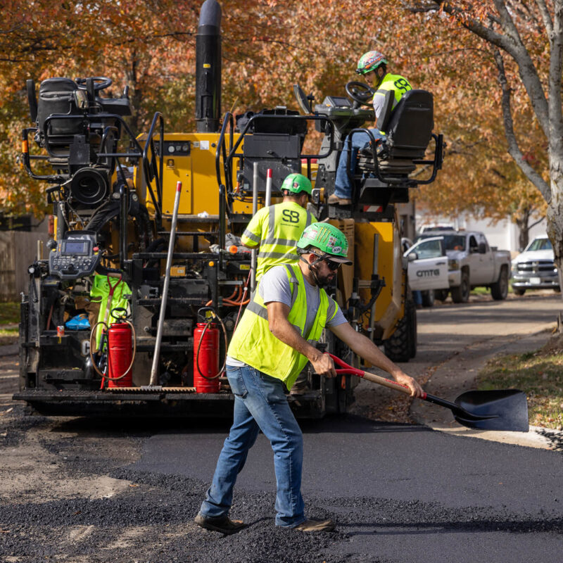 Workers repave street with asphalt; one spreads it using a shovel.