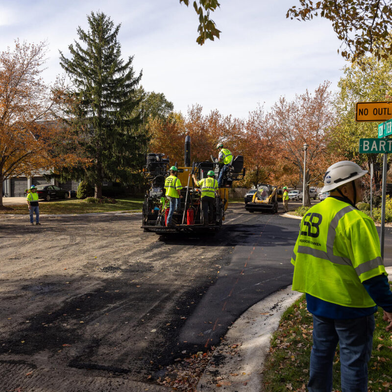 Workers repave street by Barton; machinery operates under fall trees.
