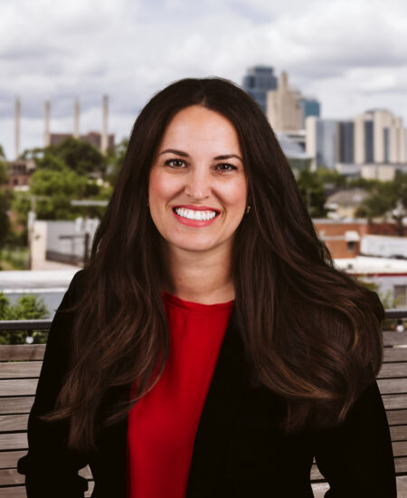 Woman in red top and black blazer smiles on outdoor bench.