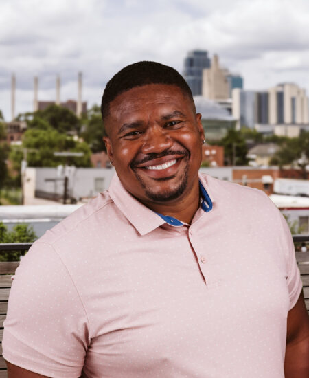 Man in pink polo smiles outdoors with city skyline behind.