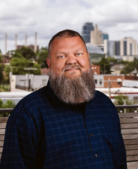 Bearded man in navy plaid shirt outdoors by city buildings.