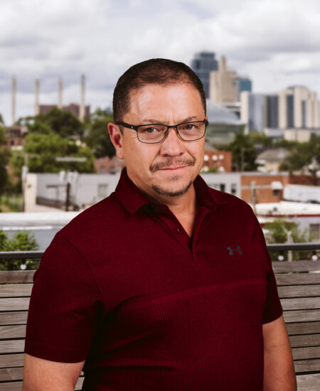 Man in glasses and maroon polo stands on outdoor bench, city behind.